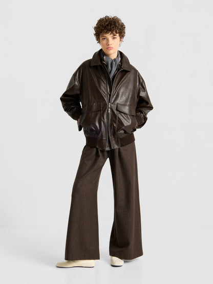 Eine Person mit kurzen Locken trägt die Oversized-Jacke aus veganem Leder in Dunkelbraun, dazu ein graues Hemd, eine braune Hose mit weitem Bein und cremefarbene Schuhe; sie steht mit den Händen in den Taschen vor einem schlichten, hellen Hintergrund.