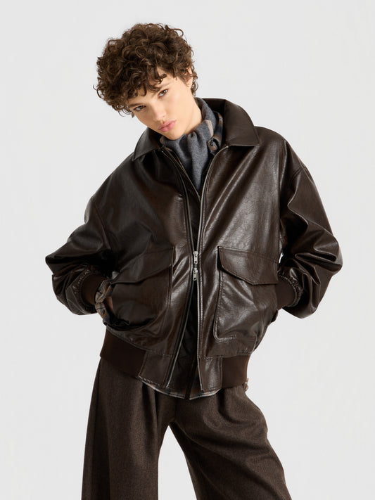 Eine Person mit kurzen Locken präsentiert „Into the Brown“: eine dunkelbraune Lederjacke über einem grauen Oberteil und einer lockeren braunen Hose, die mit den Händen in den Taschen vor einem schlichten hellen Hintergrund steht.