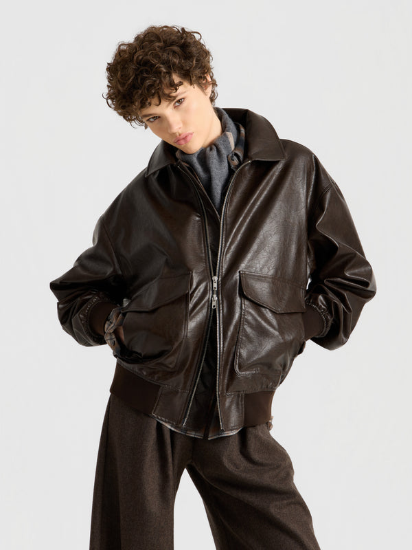 Eine Person mit kurzen Locken präsentiert „Into the Brown“: eine dunkelbraune Lederjacke über einem grauen Oberteil und einer lockeren braunen Hose, die mit den Händen in den Taschen vor einem schlichten hellen Hintergrund steht.