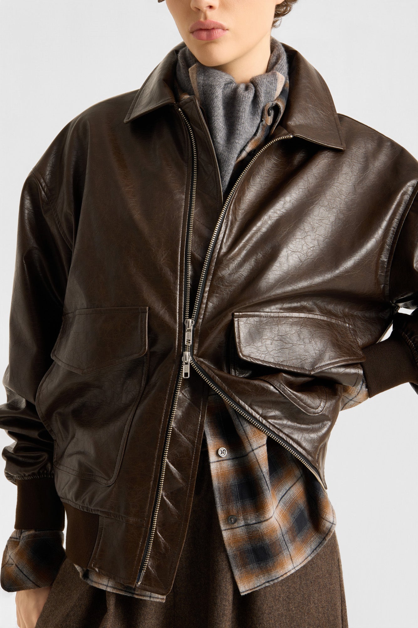 Eine Person trägt die Oversized-Jacke aus veganem Leder in Dunkelbraun über einem grauen Schal und einem karierten Hemd, dazu eine braune Hose, und steht vor einem einfarbigen Hintergrund. Nur die untere Hälfte ihres Gesichts ist zu sehen.