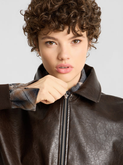 Eine Person mit lockigem Haar blickt in die Kamera, während sie eine dunkelbraune Oversize-Jacke aus veganem Leder über einem karierten Hemd zuknöpft.