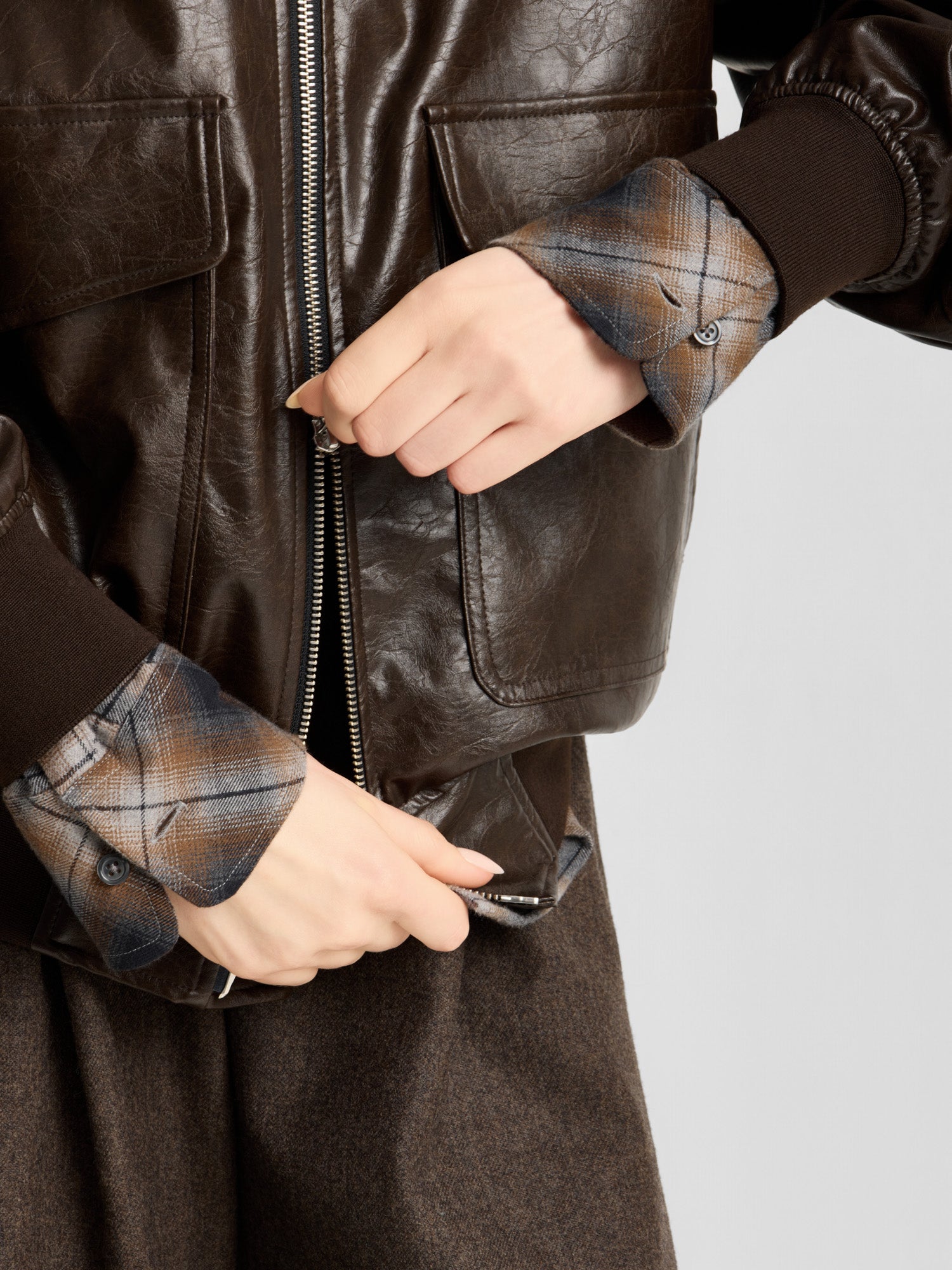 Eine Person zieht eine braune Oversize-Jacke aus veganem Leder über ein kariertes Hemd und lässt dabei eine braune Hose erkennen. Der Fokus liegt auf den Händen und den detaillierten Texturen der Kleidung.