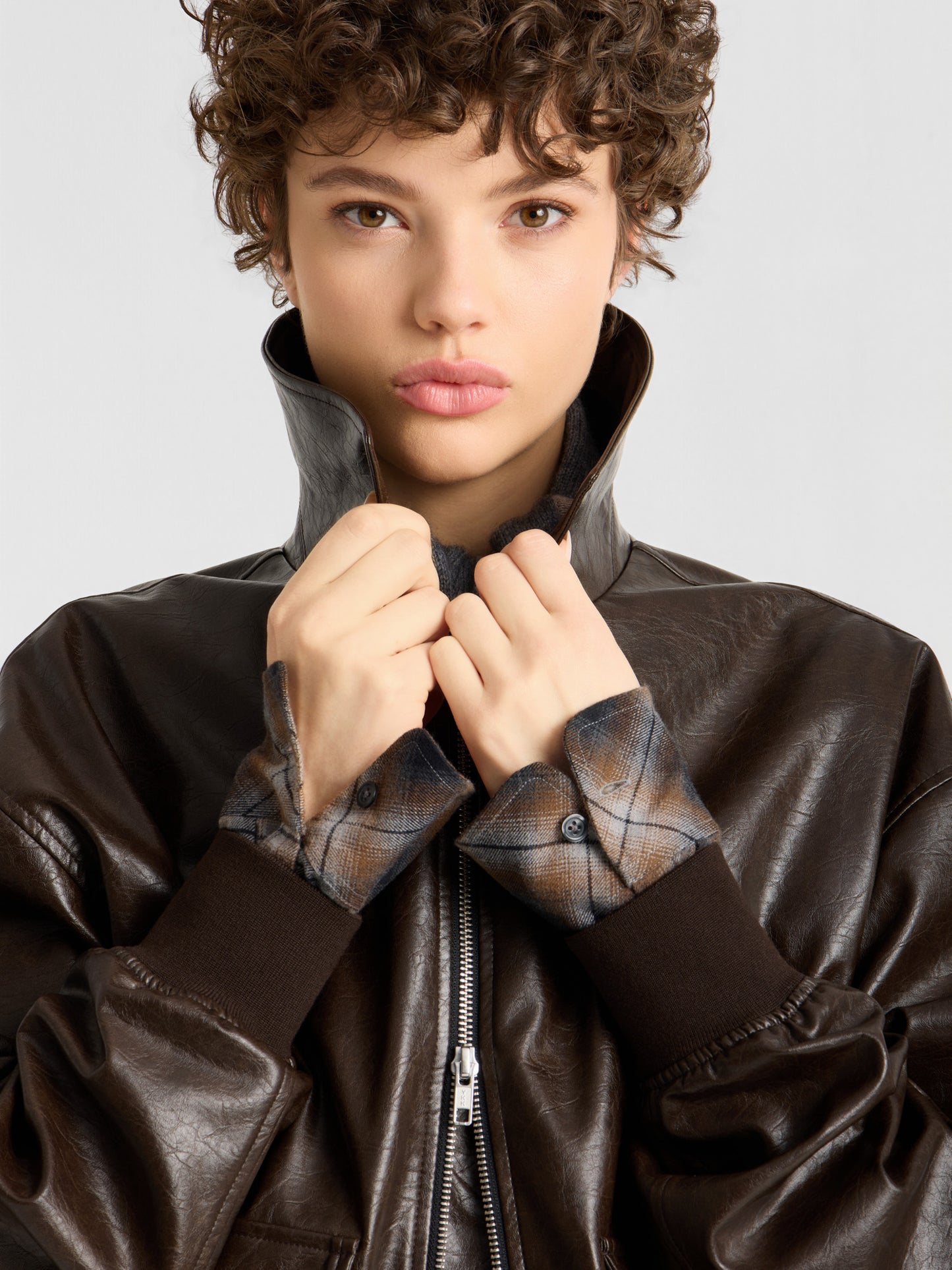 Eine Person mit kurzen Locken trägt eine Oversize-Jacke aus veganem Leder über einem karierten Hemd, hält den Kragen der Jacke mit beiden Händen hoch und blickt mit ausdruckslosem Gesicht direkt in die Kamera.