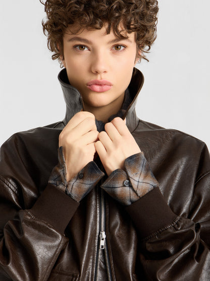 Eine Person mit kurzen Locken trägt eine Oversize-Jacke aus veganem Leder über einem karierten Hemd, hält den Kragen der Jacke mit beiden Händen hoch und blickt mit ausdruckslosem Gesicht direkt in die Kamera.