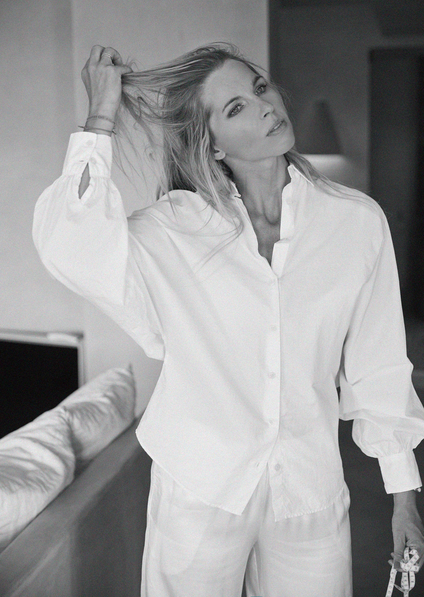 Eine Frau, die die Bluse Antonia Slim White und eine weiße Hose trägt, steht in einem Haus, hält ihr Haar mit einer Hand hoch und schaut mit entspanntem Blick in die Kamera. Das Foto ist in schwarz-weiß.