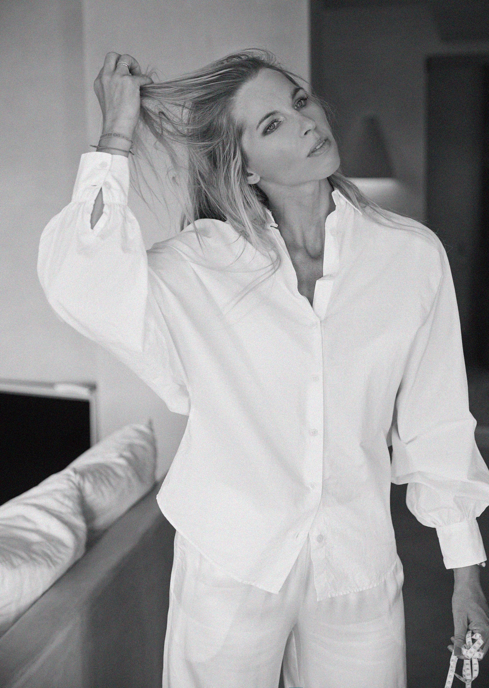 Eine Frau, die die Bluse Antonia Slim White und eine weiße Hose trägt, steht in einem Haus, hält ihr Haar mit einer Hand hoch und schaut mit entspanntem Blick in die Kamera. Das Foto ist in schwarz-weiß.
