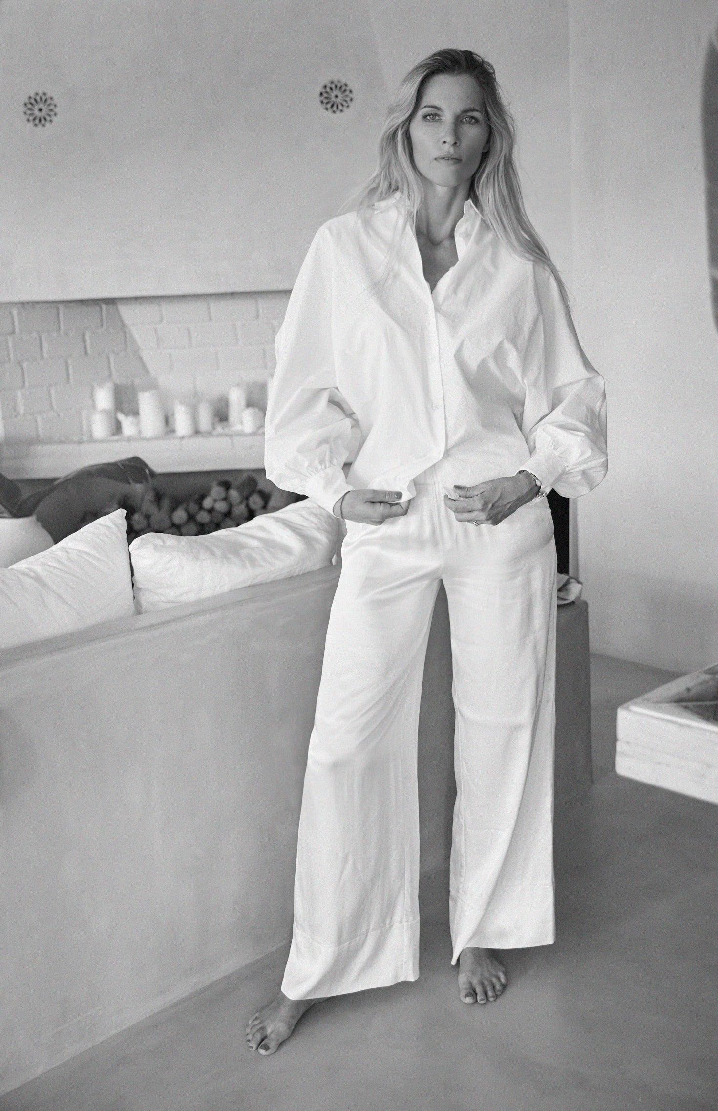 Eine Frau steht barfuß in einem Innenraum und trägt die Bluse Antonia Slim White und eine weitbeinige weiße Hose. Mit langem Haar und entspanntem Gesichtsausdruck, eine Hand in der Tasche, ergänzt sie die ruhige Atmosphäre des minimalistischen Settings.
