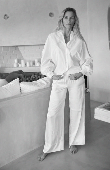 Eine Frau steht barfuß in einem Innenraum und trägt die Bluse Antonia Slim White und eine weitbeinige weiße Hose. Mit langem Haar und entspanntem Gesichtsausdruck, eine Hand in der Tasche, ergänzt sie die ruhige Atmosphäre des minimalistischen Settings.