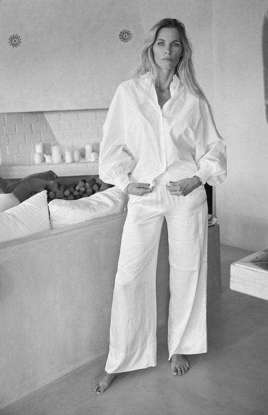 Eine Frau steht barfuß in einem Innenraum und trägt die Bluse Antonia Slim White und eine weitbeinige weiße Hose. Mit langem Haar und entspanntem Gesichtsausdruck, eine Hand in der Tasche, ergänzt sie die ruhige Atmosphäre des minimalistischen Settings.