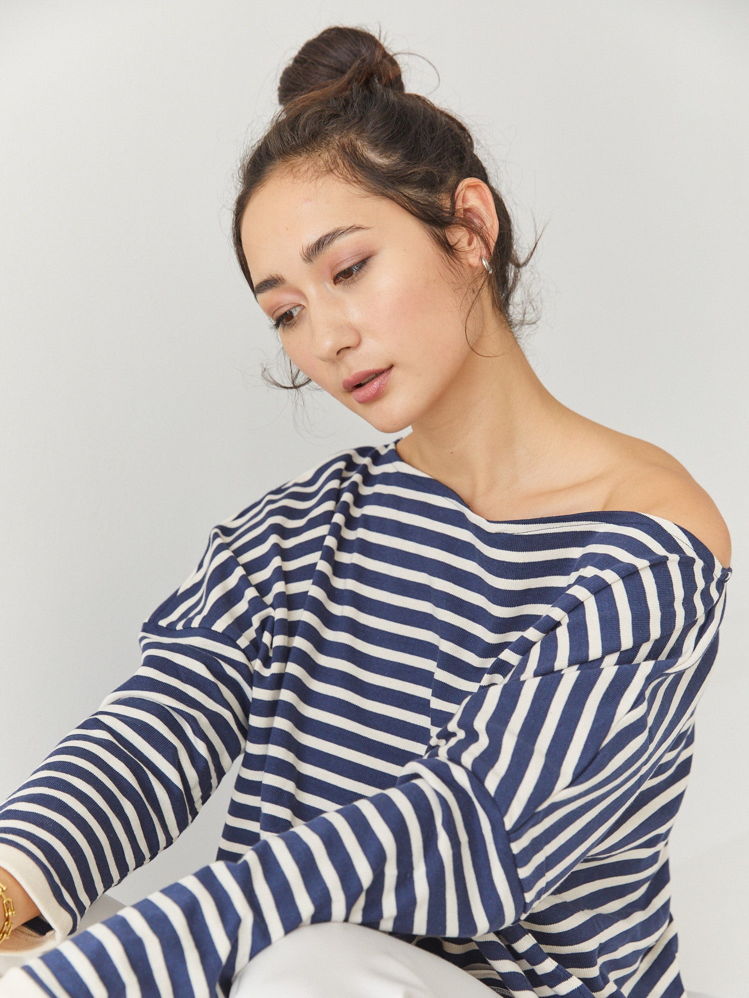 Eine Frau mit dunklem Haar in einem Dutt trägt das Sweatshirt Sailor Stripe Navy Ecru und sitzt entspannt vor einem schlichten weißen Hintergrund.