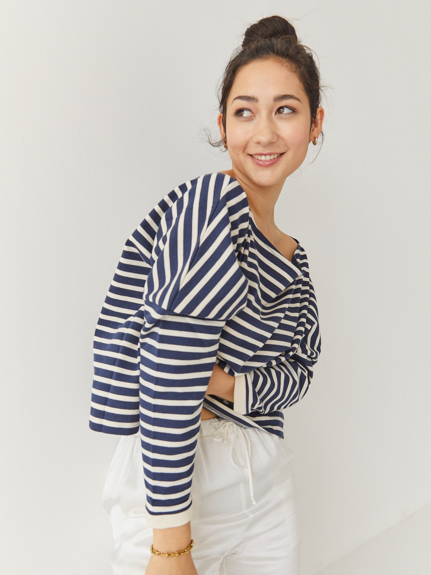 Eine lächelnde Frau mit dunklen Haaren in einem Dutt trägt das Sweatshirt Sailor Stripe Navy Ecru und eine weiße Hose und steht vor einem schlichten weißen Hintergrund.