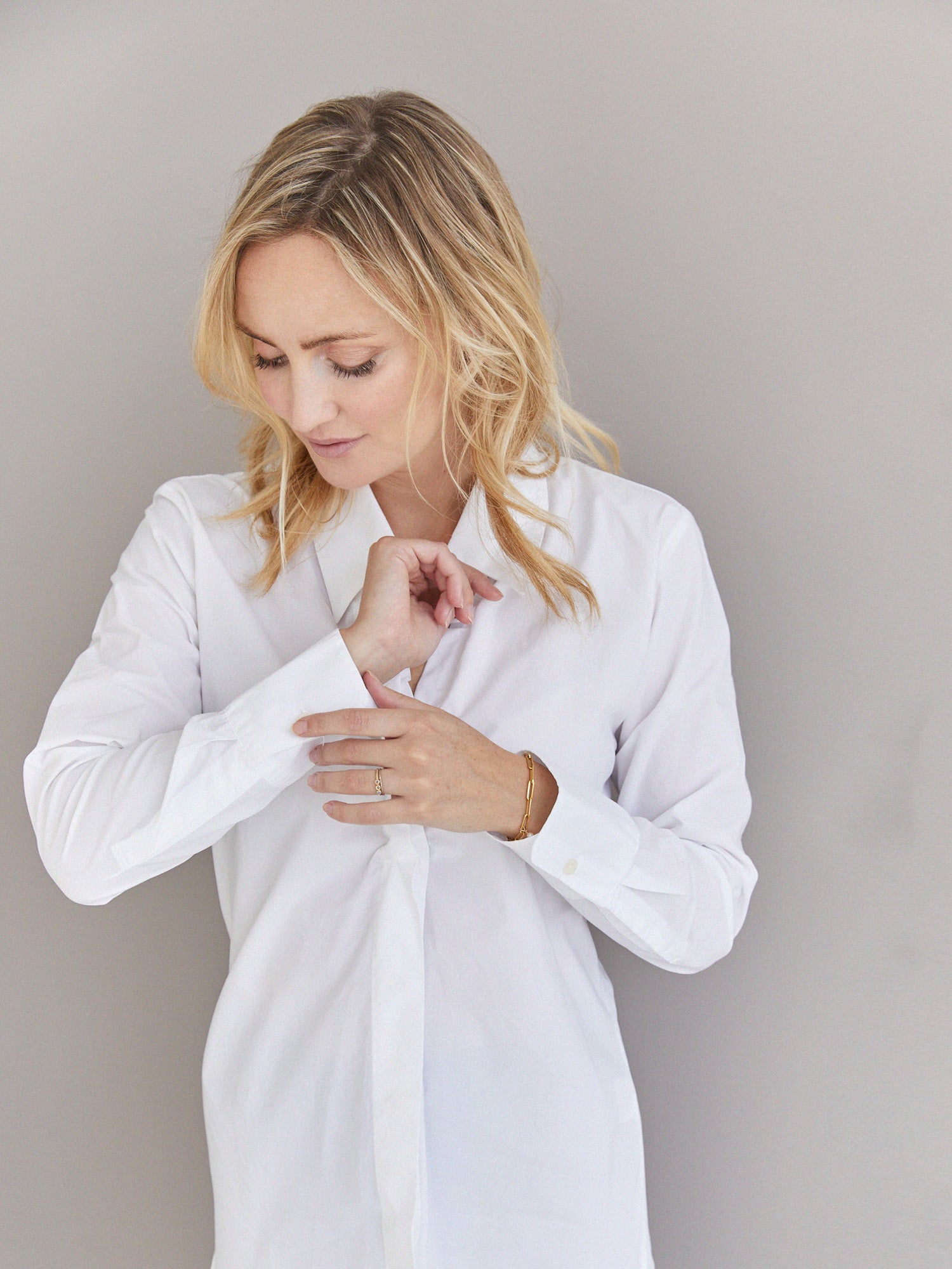 Eine blonde Frau, die die Bluse Ella White, eine taillierte weiße Baumwollbluse, trägt, schaut nach unten, während sie ihren Ärmel vor einem schlichten hellen Hintergrund zurechtrückt.