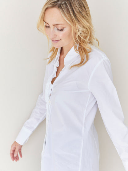 Eine Frau mit gewelltem blondem Haar trägt die taillierte Bluse Ella White aus Baumwolle und steht vor einem hellen Hintergrund. Sie blickt mit einem sanften Ausdruck nach unten.