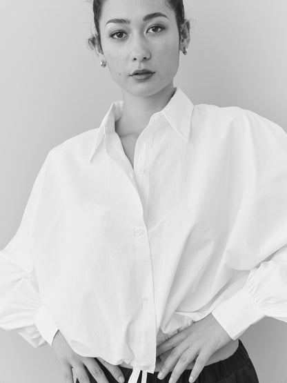 Schwarz-Weiß-Foto einer Person in locker sitzender Bluse Draw String White und dunkler Hose, stehend mit den Händen auf den Hüften vor einem schlichten Hintergrund mit einem neutralen Ausdruck.