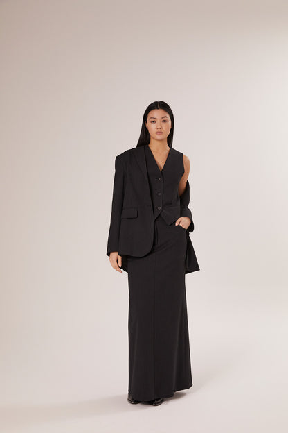 Eine Frau mit langen dunklen Haaren steht vor einem schlichten Hintergrund und trägt den Blazer Pinstripe Black als Teil eines schicken dreiteiligen Anzugs, gepaart mit einer ärmellosen Weste und einem langen Rock, selbstbewusst in die Kamera.