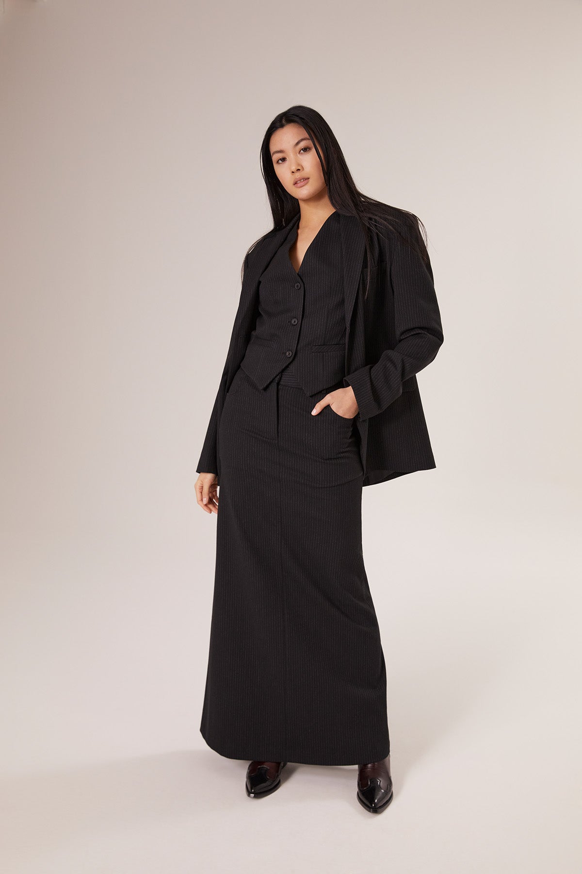 Eine Frau mit langen dunklen Haaren trägt den Blazer Pinstripe Black mit einer Weste und einem langen Rock und steht selbstbewusst vor einem schlichten hellen Hintergrund.
