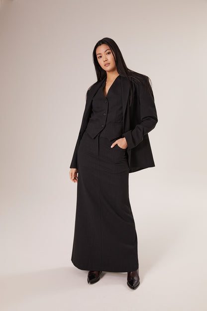 Eine Frau mit langen dunklen Haaren trägt den Blazer Pinstripe Black mit einer Weste und einem langen Rock und steht selbstbewusst vor einem schlichten hellen Hintergrund.