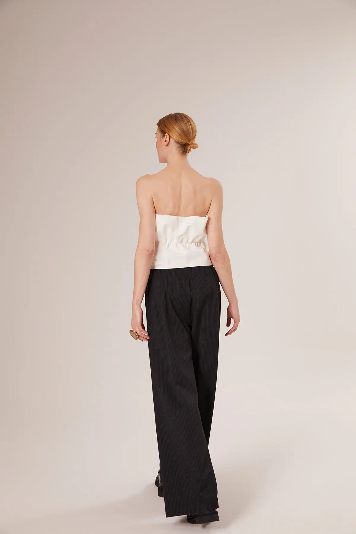 Eine Frau mit blondem Haar in einem tiefen Dutt ist von hinten zu sehen. Sie trägt ein weißes Plissee-Top und eine locker sitzende schwarze Hose und steht vor einem schlichten hellen Hintergrund.