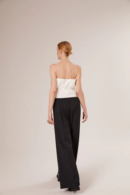 Eine Frau mit blondem Haar in einem tiefen Dutt ist von hinten zu sehen. Sie trägt ein weißes Plissee-Top und eine locker sitzende schwarze Hose und steht vor einem schlichten hellen Hintergrund.