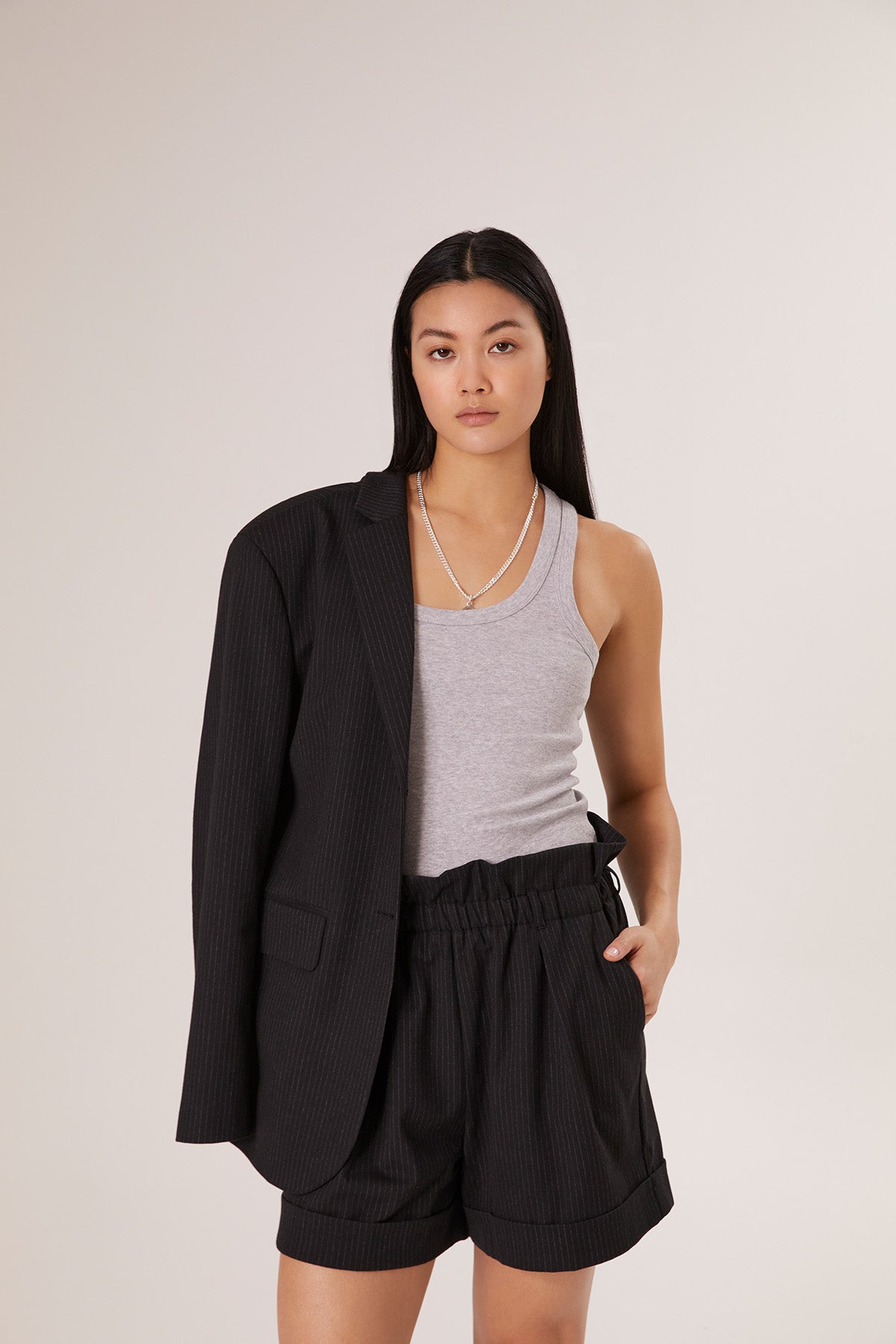 Eine Frau mit langen schwarzen Haaren trägt das Tank Top Rip Grey, schwarze Nadelstreifen-Shorts und einen passenden Blazer, der über eine Schulter drapiert ist. Sie präsentiert nachhaltige Frauenmode und steht selbstbewusst vor einem schlichten, hellen Hintergrund.