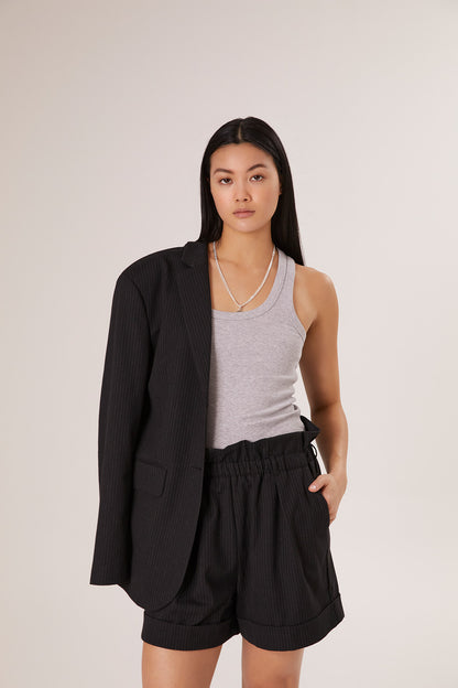 Eine Frau mit langen schwarzen Haaren trägt das Tank Top Rip Grey, schwarze Nadelstreifen-Shorts und einen passenden Blazer, der über eine Schulter drapiert ist. Sie präsentiert nachhaltige Frauenmode und steht selbstbewusst vor einem schlichten, hellen Hintergrund.