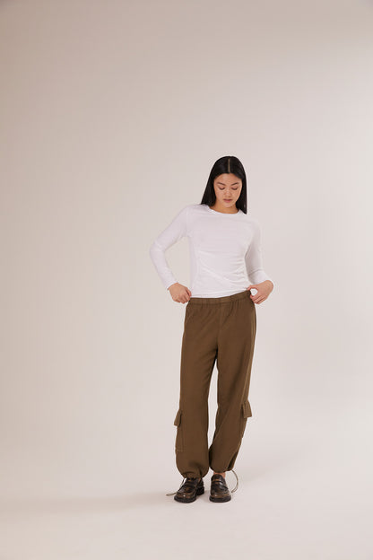 Eine Frau mit langen dunklen Haaren trägt das Hemd Gathering White und eine lockere olivgrüne Cargohose. Sie steht vor einem schlichten hellen Hintergrund und hat die Hände in den Taschen - ein stilvolles Beispiel für nachhaltige Mode.