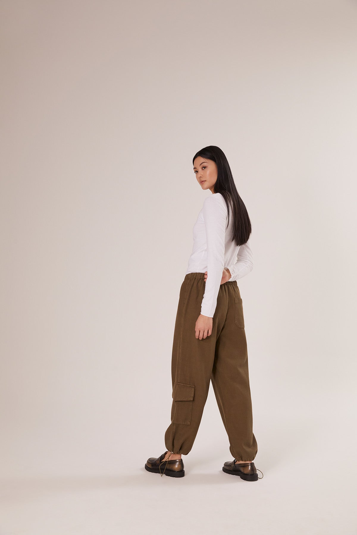 Eine Frau steht in einem minimalistischen Studio und trägt das Shirt Gathering White, gepaart mit einer lockeren braunen Cargohose und schwarzen Schuhen, und blickt über ihre Schulter - ein neutraler, nachhaltiger Mode-Look.