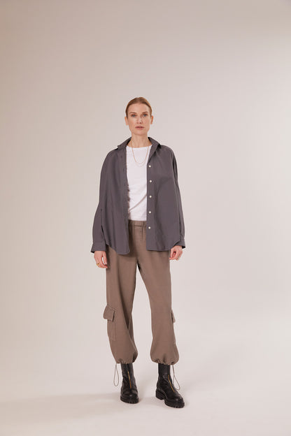 Eine Frau blickt nach vorne, sie trägt eine Bluse Boyfriend Anthrazit über einem weißen T-Shirt, dazu eine taupefarbene Cargohose und schwarze Ankle Boots vor einem schlichten hellen Hintergrund.