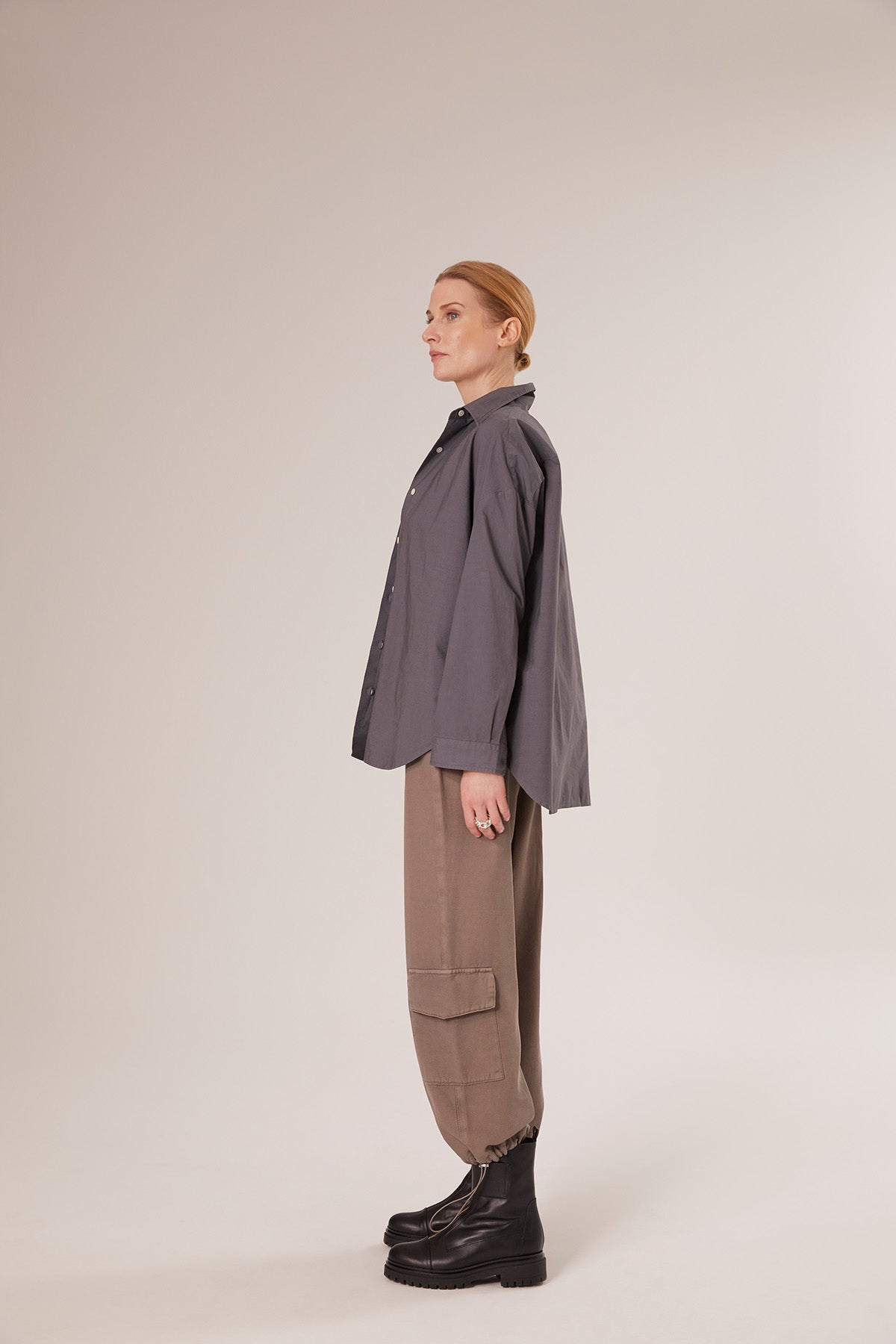 Eine Person mit blondem Haar in einem niedrigen Dutt steht im Profil und trägt einen Bluse Boyfriend Anthrazit, eine hellbraune Cargohose und schwarze Stiefel vor einem schlichten hellen Hintergrund.