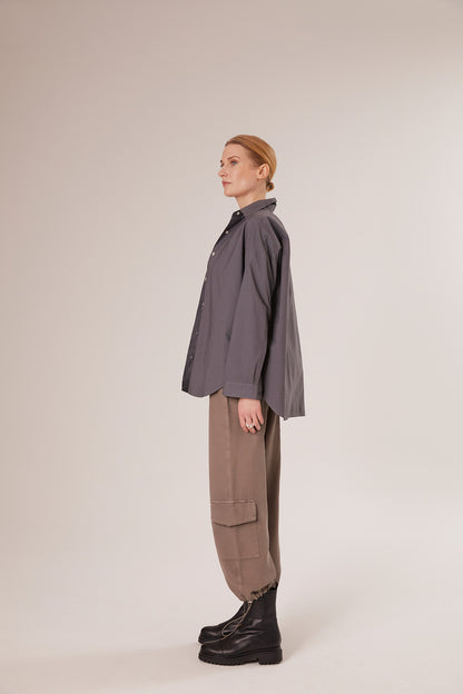 Eine Person mit blondem Haar in einem niedrigen Dutt steht im Profil und trägt einen Bluse Boyfriend Anthrazit, eine hellbraune Cargohose und schwarze Stiefel vor einem schlichten hellen Hintergrund.