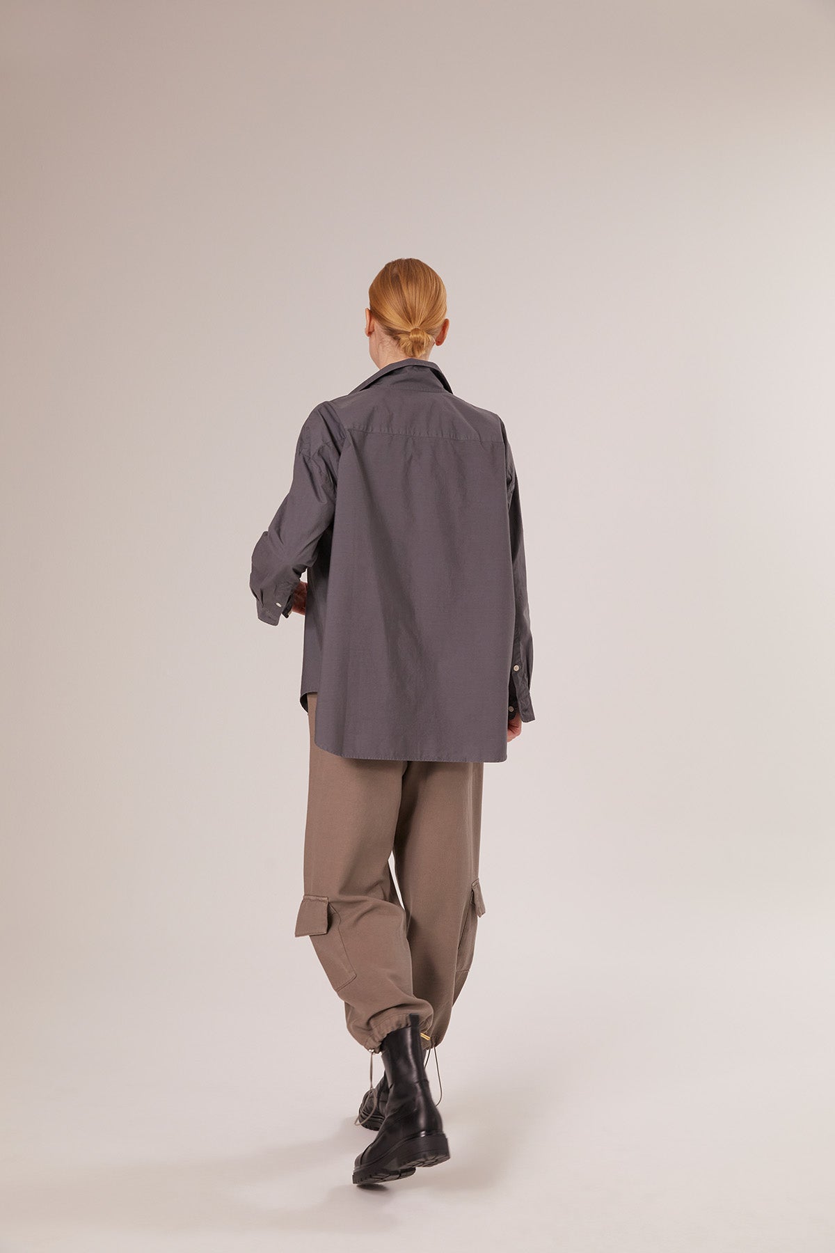 Eine Person mit blondem Haar, bekleidet mit einem grauen Bluse Boyfriend Anthrazit, einer taupefarbenen Hose mit Seitentaschen und schwarzen Stiefeln, geht in einem minimalistischen Studio von der Kamera weg.