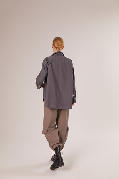 Eine Person mit blondem Haar, bekleidet mit einem grauen Bluse Boyfriend Anthrazit, einer taupefarbenen Hose mit Seitentaschen und schwarzen Stiefeln, geht in einem minimalistischen Studio von der Kamera weg.