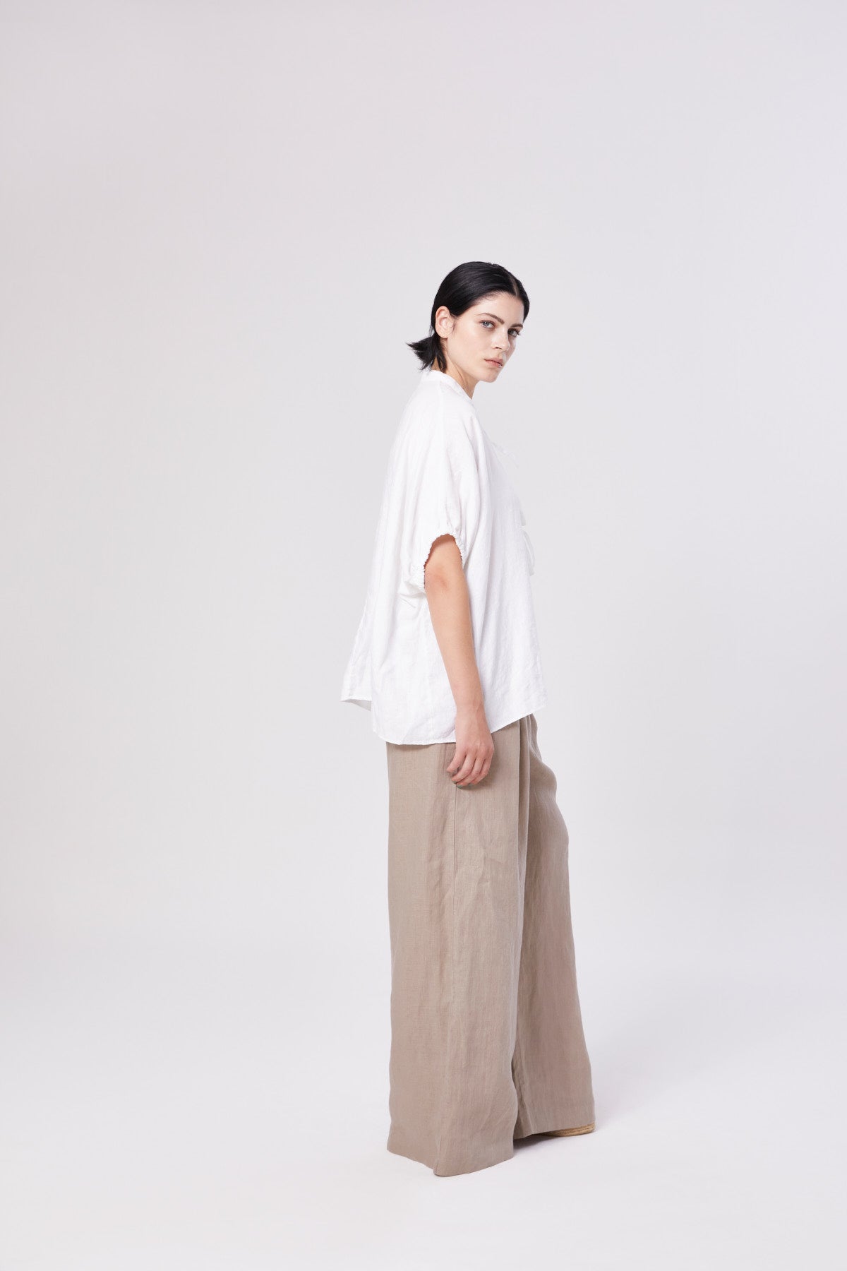 Eine Person mit kurzen dunklen Haaren steht seitlich, trägt die Bluse Cowboy Bow und eine beigefarbene Hose mit weiten Beinen, die die nachhaltige Mode vor einem schlichten hellen Hintergrund umarmt.