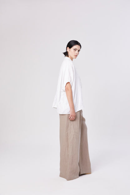 Eine Person mit kurzen dunklen Haaren steht seitlich, trägt die Bluse Cowboy Bow und eine beigefarbene Hose mit weiten Beinen, die die nachhaltige Mode vor einem schlichten hellen Hintergrund umarmt.
