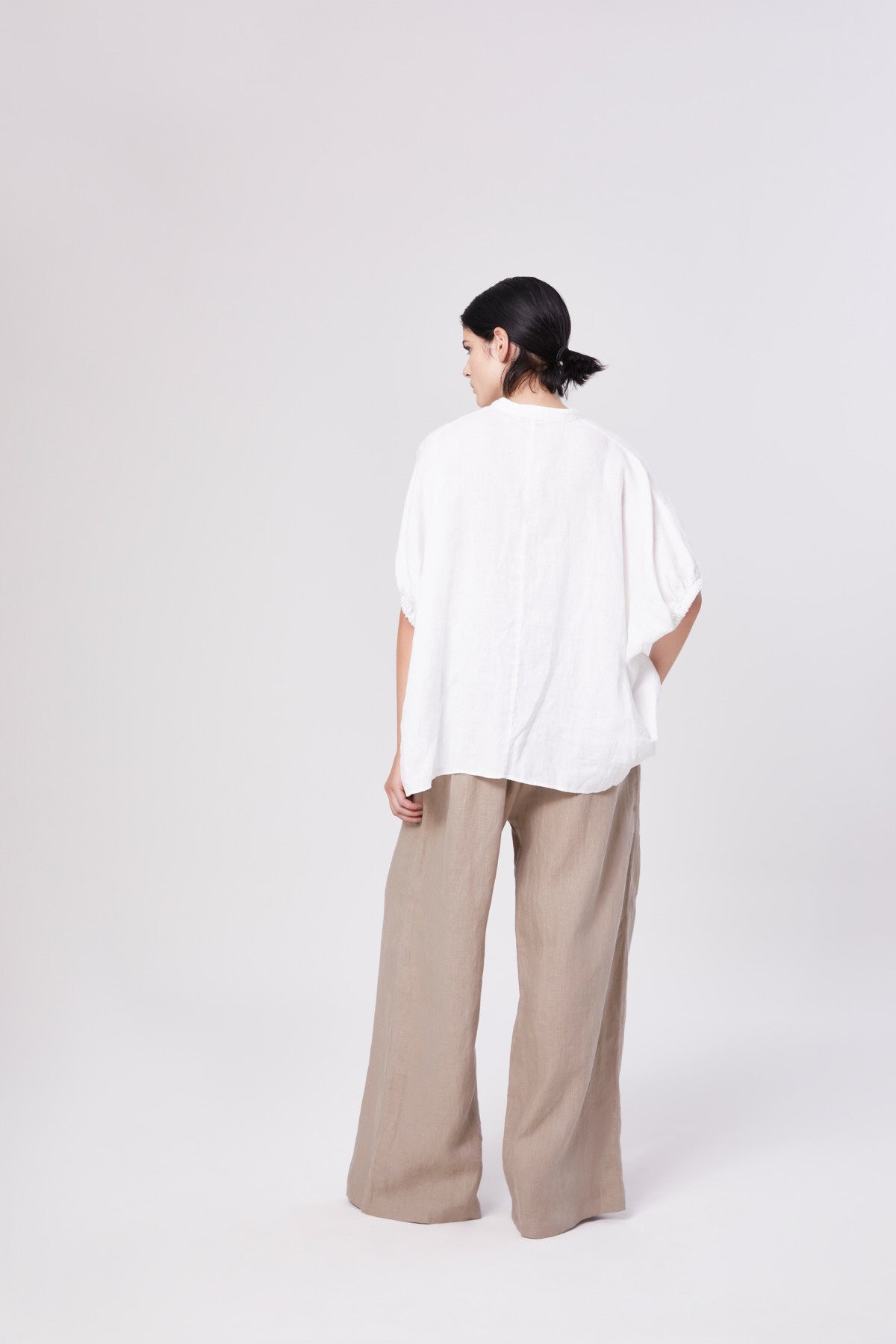 Eine Person mit dunklen Haaren in einem tiefen Dutt steht mit dem Blick zur Seite, trägt die Bluse Cowboy Bow und eine beigefarbene Hose mit weitem Bein - ein müheloser Look, der nachhaltige Mode vor einem schlichten hellen Hintergrund verkörpert.