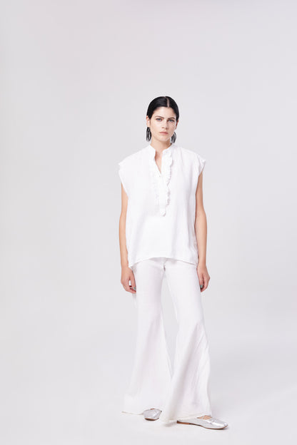 Eine Frau steht vor einem schlichten weißen Hintergrund und trägt eine lockere weiße kurzärmelige Bluse mit Knöpfen, die Hose Joana White, silberne Schuhe und schwarze Ohrringe.