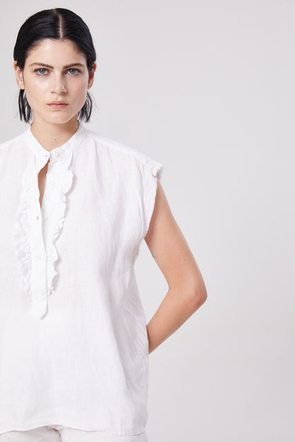 Eine Person mit dunklem, nach hinten gestyltem Haar, die eine ärmellose weiße Rüschenbluse und die Hose Joana White trägt, steht mit einem Arm hinter dem Rücken vor einem schlichten hellen Hintergrund.