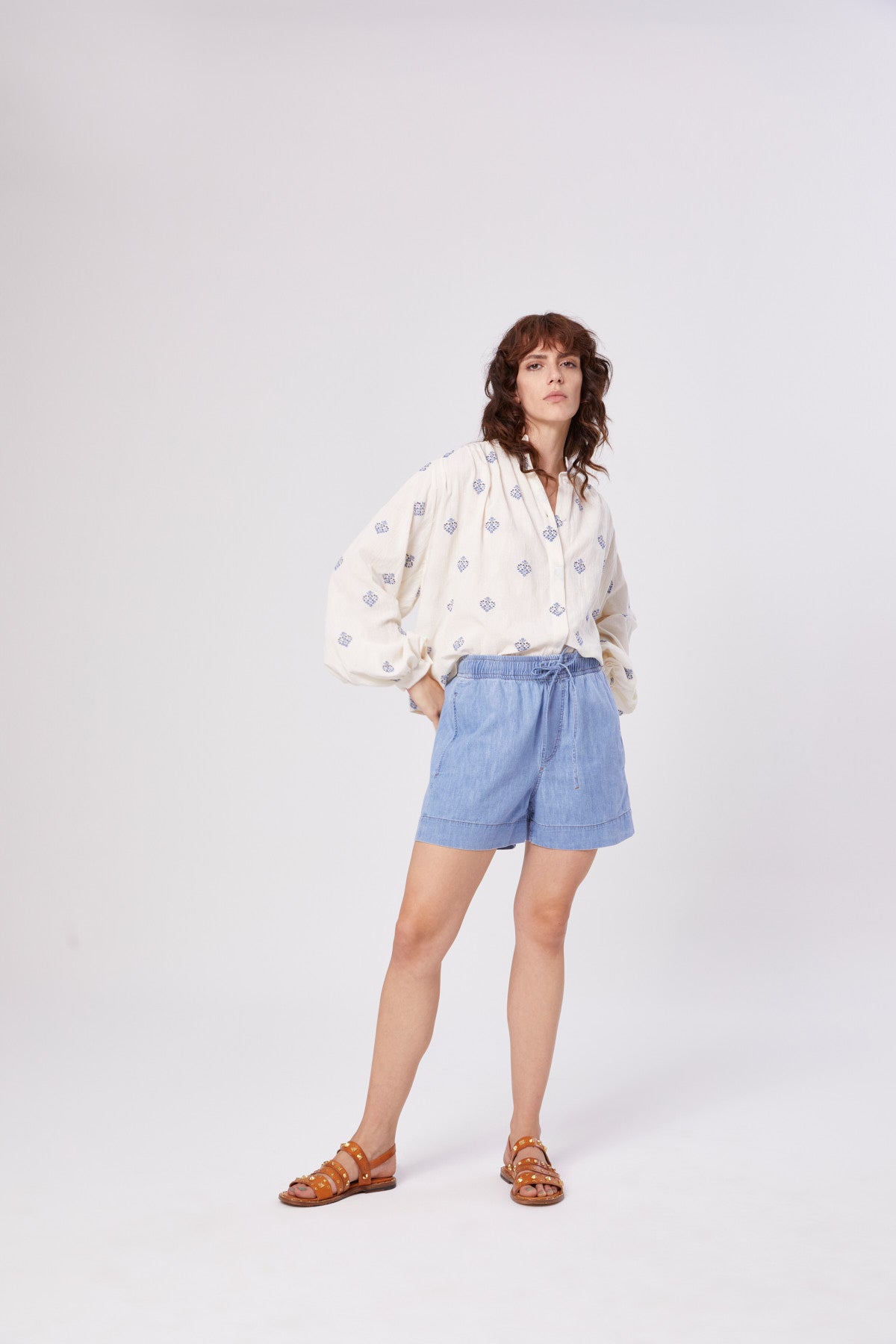 Eine Frau mit gewelltem braunem Haar steht vor einem weißen Hintergrund und trägt eine kurze Jeans, eine weite weiße Bluse mit blauen Mustern und braune Sandalen. Sie hat eine entspannte Körperhaltung und eine Hand in der Tasche.