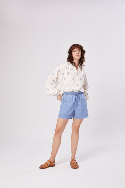 Eine Frau mit gewelltem braunem Haar und entspanntem Gesichtsausdruck steht vor einem schlichten weißen Hintergrund und trägt eine gemusterte weiße Bluse, braune Sandalen und die Shorts Light Denim in Hellblau mit Tunnelzug.