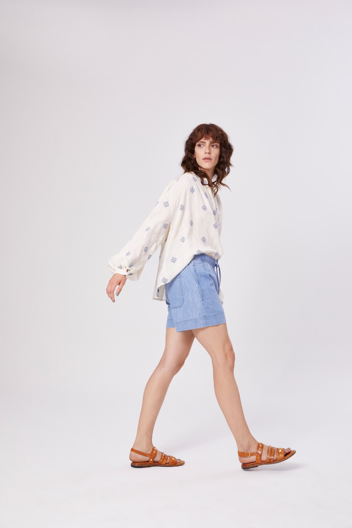 Eine Frau mit gewelltem braunem Haar geht im Profil vor einem weißen Hintergrund, trägt die Bluse-Stickerei, hellblaue Shorts und braune Sandalen.