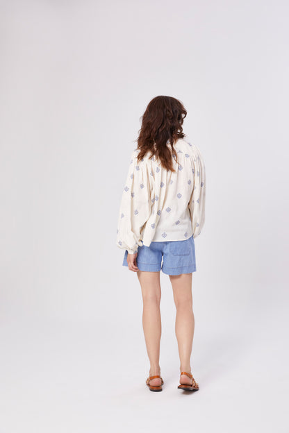 Eine Person mit gewelltem braunem Haar, in einer lockeren weißen langärmeligen Bluse mit blauem Muster, Shorts aus heller Jeans und braunen Sandalen steht mit dem Gesicht zur Seite auf einem schlichten hellen Hintergrund.