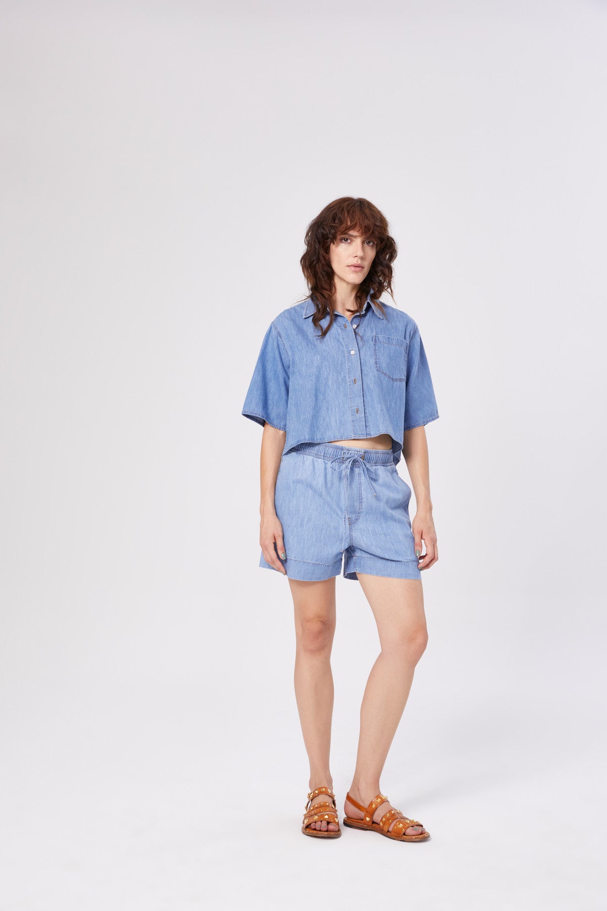 Eine Person mit gewelltem braunem Haar trägt die Bluse Short Sleeve Denim in Hellblau mit passenden Shorts mit Kordelzug und braunen Sandalen vor einem schlichten hellen Hintergrund.