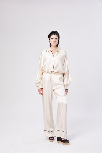 Eine Frau steht vor einem weißen Hintergrund und trägt das Bluse Antonia PJ-Set in Cremeweiß, mit langen Ärmeln und einer Hose mit weitem Bein für einen eleganten Look. Sie kombiniert es mit schwarzen Plateausandalen und frisiert ihr dunkles Haar nach hinten.