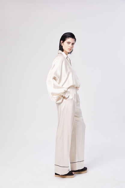 Eine Person mit dunklen Haaren modelliert die Bluse Antonia PJ in Cremeweiß mit passender Hose mit weitem Bein. Sie steht in seitlicher Pose vor einem weißen Hintergrund, um den eleganten damenbluse-Stil zu betonen.