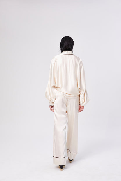 Eine Person mit dunklem Haar, von hinten gesehen, trägt eine cremefarbene Pants PJ und eine passende Bluse mit schwarzem Saum - ein eleganter, an einen Pyjama erinnernder Look - und steht vor einem schlichten weißen Hintergrund.