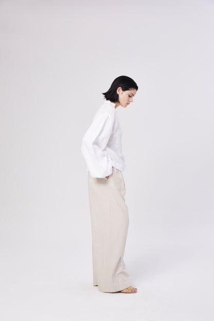 Eine Person mit kurzen dunklen Haaren, die das Bluse Top Antonia Linen mit voluminösen Ärmeln und eine beigefarbene Hose mit weitem Bein trägt, steht im Profil vor einem schlichten weißen Hintergrund.