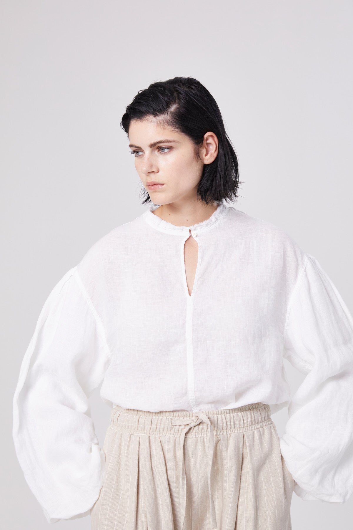 Eine Person mit kurzen dunklen Haaren trägt das Bluse Top Antonia Linen in Weiß mit voluminösen Ärmeln und eine beigefarbene, geraffte Hose mit hoher Taille. Sie steht vor einem schlichten hellen Hintergrund und schaut leicht zur Seite.