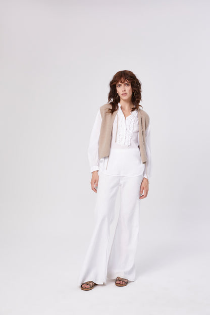 Eine Frau steht vor weißem Hintergrund und trägt die Bluse Bruna white mit Rüschen, eine cremefarbene Strickweste, weite weiße Hosen und braune Sandalen. Sie hat welliges braunes Haar und einen neutralen Gesichtsausdruck.