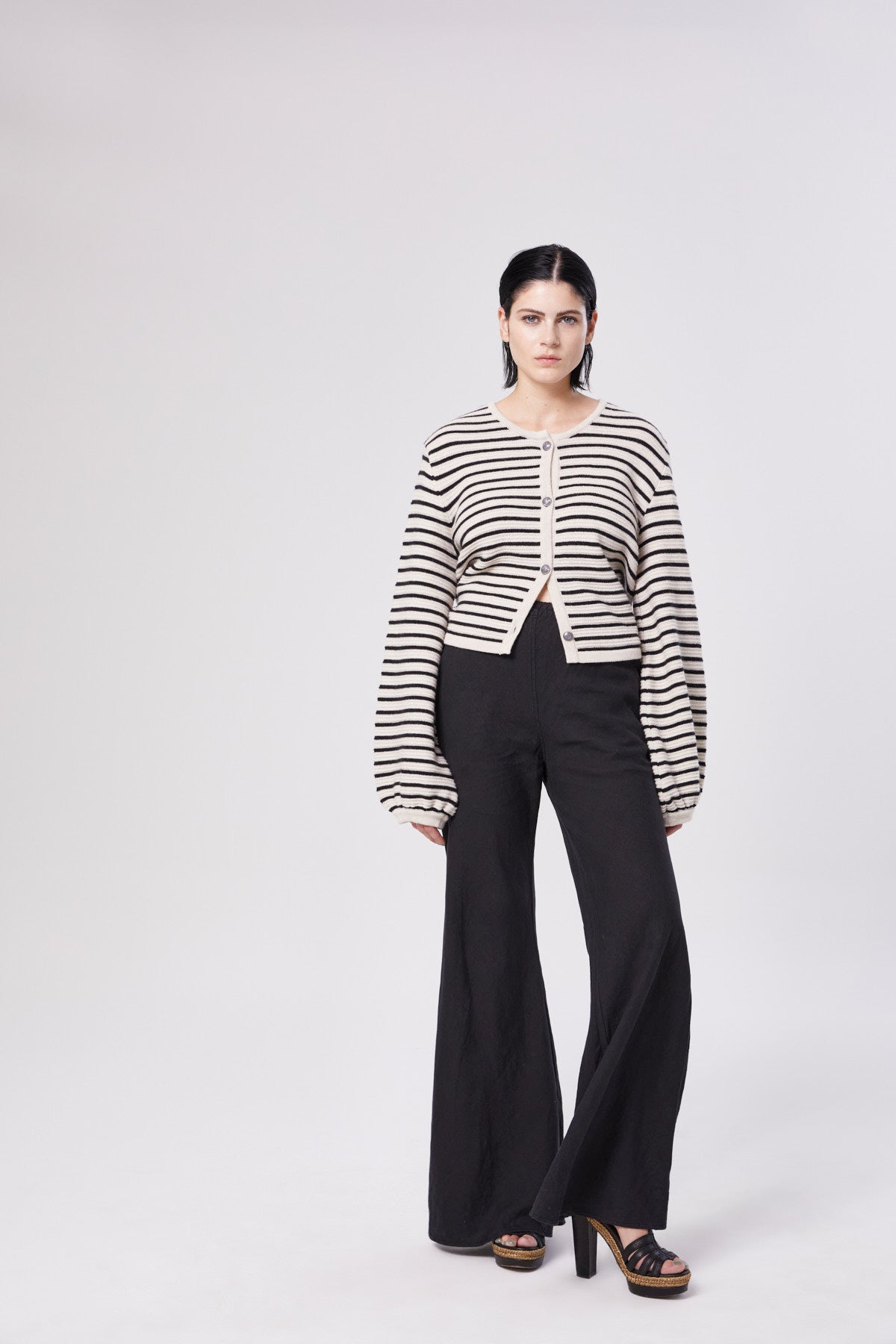 Eine Frau mit glattem dunklem Haar steht vor einem schlichten Hintergrund und trägt die übergroße Jacke Fiene mit beigen und schwarzen Streifen und weiten Ärmeln, gepaart mit einer schwarzen Hose mit weitem Bein und schwarzen Plateauabsätzen.