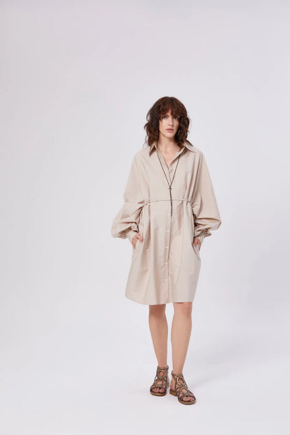 Eine Person mit schulterlangem braunem Haar trägt das Dress Antonia Dress Vol. 2 Beige, ein langärmeliges Hemdkleid mit Taschen. Sie steht vor einem schlichten weißen Hintergrund und trägt braune Sandalen.